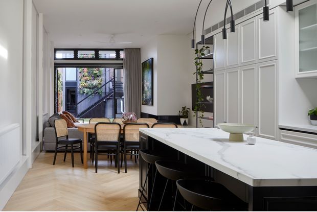 A bright, refined open-plan kitchen and dining area. The foreground features a large white marble-look island with dark cabinetry and black bar stools, while the background includes a timber dining table, woven-back chairs, a beige sofa, white decorative wall panelling, black-framed glazing, and arched pendant lighting. Light timber flooring and abundant daylight create an inviting contemporary interior.