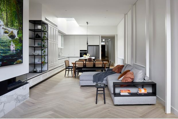 A bright, open-plan contemporary living and dining area with a large grey sectional sofa, black coffee table, timber dining table and chairs, and extensive white built-in cabinetry. The room features pale herringbone timber flooring, white walls and ceiling, black open shelving, large artwork, and a marble-look fireplace surround or media wall.