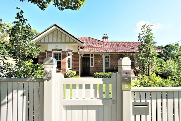 Front exterior view of a single-storey brick bungalow with a red tiled gable roof, prominent timber-framed gable detailing, white-painted picket fencing, and a gated entrance. Mature garden planting surrounds the house, with the facade and front boundary forming the main subject.