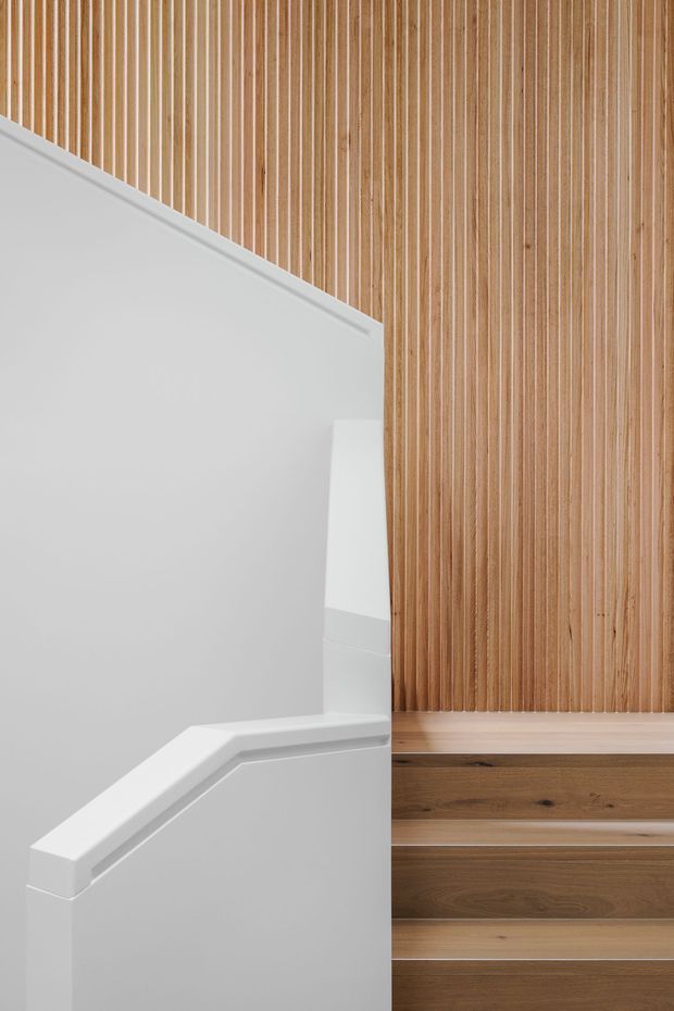 Close-up architectural view of a minimalist stairway. White painted walls and a sculptural white handrail contrast with timber stair treads, risers, and a full-height vertical timber slat wall. The composition emphasizes clean lines, warm wood texture, and a crisp neutral palette.