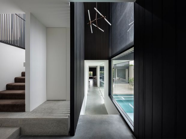 A dramatic contemporary interior hallway or landing with polished concrete flooring, white walls, dark vertical slatted feature walls and a large glazed opening overlooking an indoor pool. A carpeted stairway rises on the left, while a sculptural linear pendant hangs in the tall void above. The space is minimalist, monochromatic and strongly architectural, with glimpses of adjoining living areas beyond.