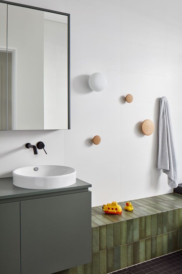 Contemporary bathroom featuring a floating dark grey vanity with a white vessel basin, wall-mounted black tapware, a large black-framed mirrored cabinet, and a tiled bathtub surround in muted green rectangular tiles. White wall tiles, rounded wooden wall hooks, a grey towel, and playful yellow bath toys add warmth and character.