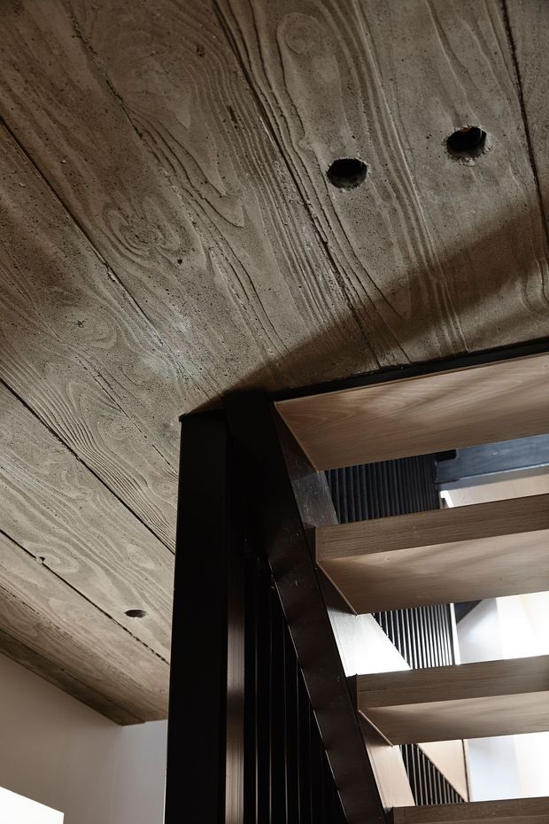 Close-up architectural interior detail looking upward at a contemporary stairway. Broad exposed concrete ceiling slabs with visible timber-like formwork grain and recessed dark openings are supported by dark structural steel or timber framing. A flight of warm timber stair treads with open risers and a black vertical slatted balustrade rises through a bright stair void.