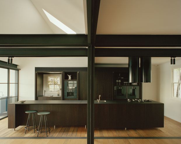 A sophisticated open-plan kitchen viewed from the living area, featuring dark timber cabinetry and a large central island with two stools. Strong black steel structural beams and posts frame the space, while exposed ductwork, linear track lighting, timber flooring, white walls, and high ceilings create a refined industrial character. A skylight or clerestory opening brings daylight into the room, and glazing is visible to the left and right.