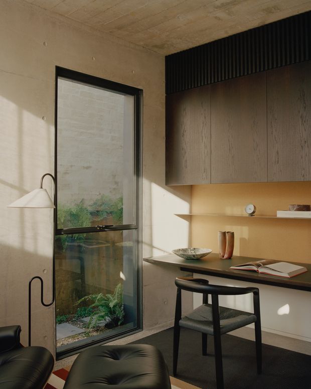 A compact, refined home office or study with a built-in timber desk and overhead cabinetry, exposed concrete walls and ceiling, dark-framed full-height glazing, and a black sculptural wall lamp. Warm sunlight enters from the window, illuminating a small landscaped lightwell or courtyard outside. The room includes a dark upholstered chair, ottoman, open book, ceramic objects, and minimalist shelving.