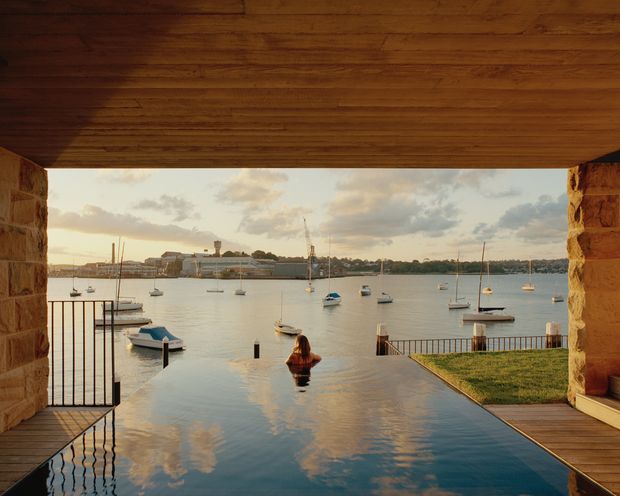 A dramatic waterfront view framed by a timber-lined ceiling and rough stone walls, looking from an inundated terrace or pool toward a marina with moored boats and a distant shoreline. The reflective water reaches into the foreground, with a person standing in it and a small grass lawn to the right. Warm, natural materials and golden late-day light create a tranquil architectural setting.