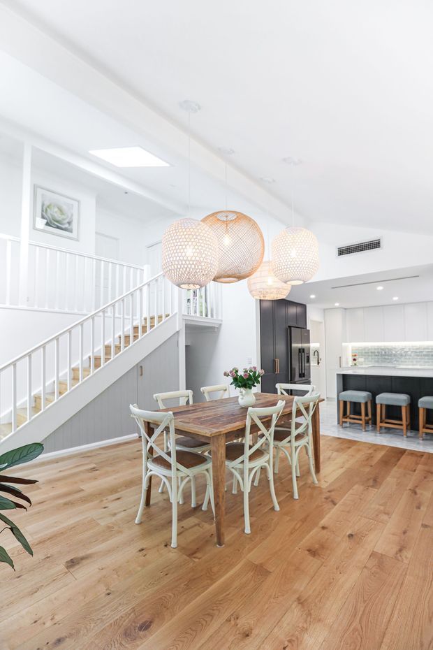 Bright open-plan dining and kitchen interior with a warm timber dining table and light woven chairs beneath multiple woven pendant lights. White walls, cabinetry, and stair balustrade create an airy coastal feel, while a dark kitchen feature wall and island provide contrast. The room opens directly to a staircase and includes light timber flooring, a tiled backsplash, and a breakfast-bar area.