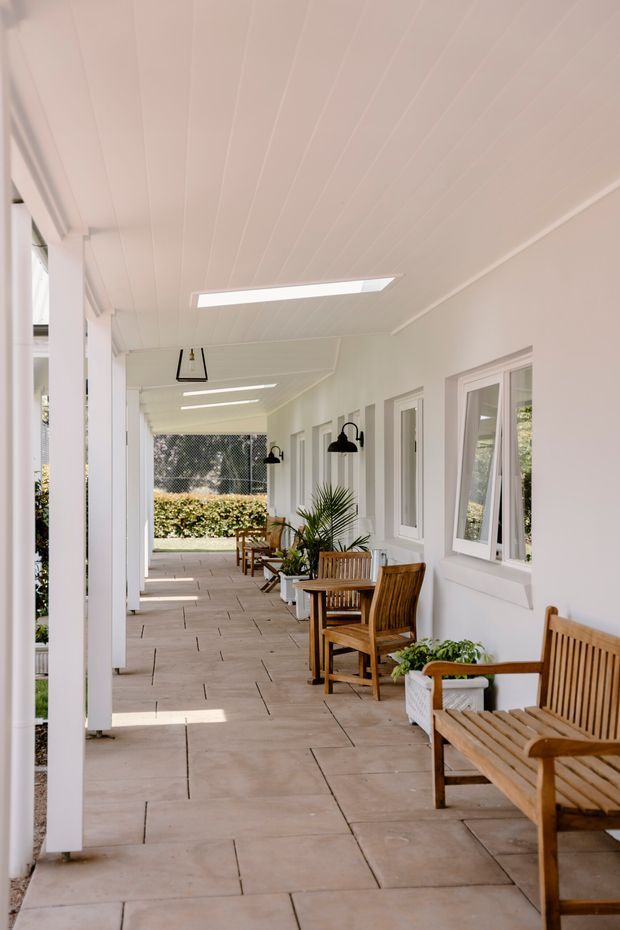 A bright, covered outdoor veranda or gallery walkway with white-painted posts, walls, windows, and a paneled ceiling. Large rectangular stone-look floor tiles run along the corridor, which is furnished with timber slatted chairs and potted plants. Black wall-mounted lanterns and linear ceiling lights provide illumination, while greenery is visible at the open end.