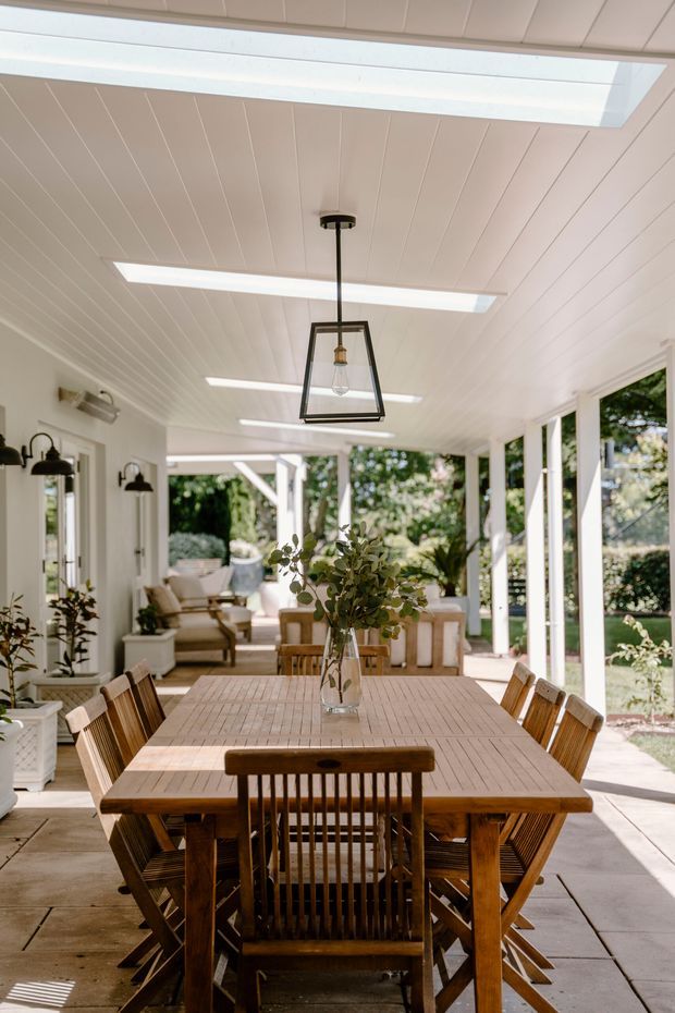 Bright covered outdoor dining area with a large solid timber dining table and matching wooden chairs, white beadboard-style ceiling, multiple skylights, black-framed pendant lighting, and extensive white-framed glazing opening onto a garden. A relaxed seating area and abundant greenery are visible beyond the dining zone.