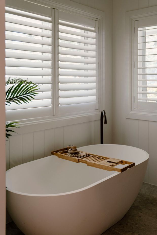 A serene, light-filled bathroom featuring a freestanding oval bathtub, wall-mounted dark tapware, white plantation shutters, and white vertical wall panelling. A timber bath caddy spans the tub, with greenery visible at the edge of the frame. The palette is soft white and warm neutral with natural timber accents.
