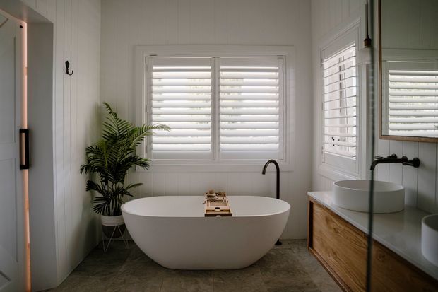 Bright, refined bathroom featuring a freestanding oval bathtub beneath plantation-shutter windows, white vertical wall panelling, a dark curved floor-mounted tap, and a timber vanity with vessel basins and large mirrored cabinetry. A potted fern adds a natural accent.