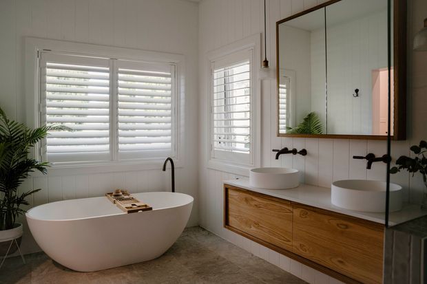 Bright, refined bathroom featuring a freestanding white oval bathtub, twin vessel basins, a floating timber vanity, black wall-mounted tapware, large framed mirror, white vertical wall panelling, plantation shutters, and a stone-look floor. Natural greenery adds an organic accent.