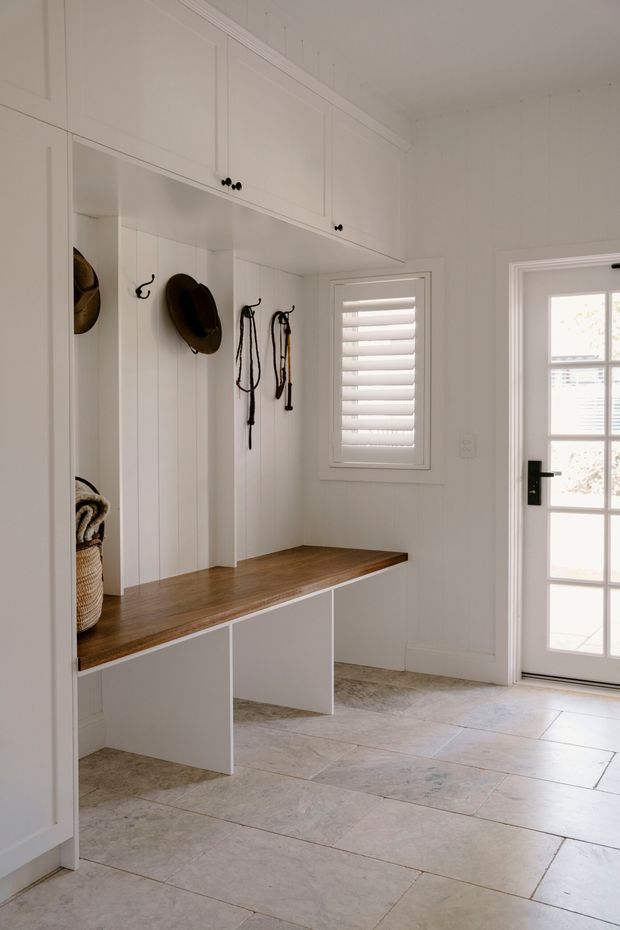 Bright, refined entryway or mudroom with custom white built-in cabinetry, open cubbies, hooks, upper storage cupboards, and a warm timber bench seat. The room has white vertical wall panelling and trim, a shuttered window, glazed exterior door, and large pale stone-look floor tiles.