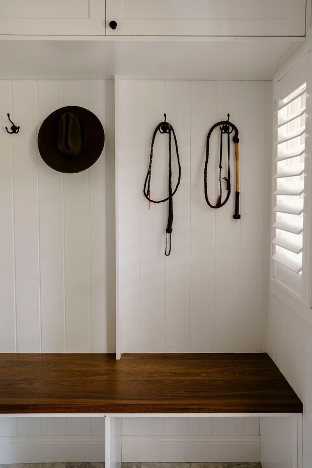 A compact white paneled entryway or mudroom with built-in overhead cabinetry, a dark timber bench seat, black hooks, and equestrian-style bridles and a hat mounted on the wall. White plantation shutters provide filtered daylight on the right, while the timber bench and dark wood floor add warmth.
