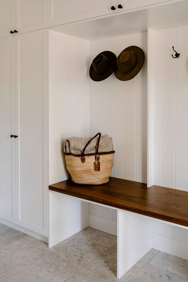 Built-in entryway mudroom storage with white paneled cabinetry, an integrated dark timber bench, vertical wall panelling, hooks, and open cubbies. A woven basket and hats add warm rustic detail, while pale stone-look floor tiles create a light, practical finish.