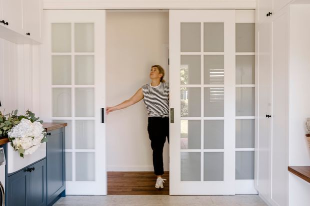 Bright, refined interior view of a white built-in cabinetry-lined passage with a pair of white French-style glazed pocket or sliding doors opening onto a timber-floored room. Dark blue lower cabinetry and timber benchtops are visible at left and right, with a floral arrangement and a person entering through the doorway.
