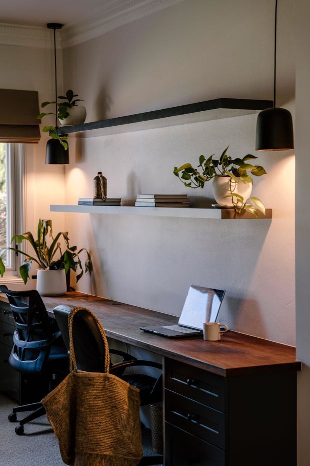 A compact, warmly lit home office with a long dark timber desk, black drawer cabinetry, two black task chairs, wall-mounted floating shelves, pendant lights, indoor plants, and a laptop. Decorative ceiling cornices and a muted neutral palette add a refined traditional architectural detail to the otherwise contemporary workspace.