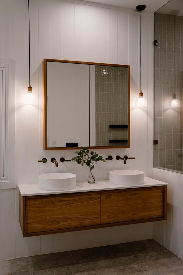 A refined bathroom with a floating double vanity, two round white vessel basins, wall-mounted dark metal tapware, and warm timber cabinetry. A large timber-framed mirrored cabinet hangs on white vertically panelled walls, with a tiled shower or bath area visible at right. Pendant lights flank the mirror, and the floor has a dark mottled stone-like finish.