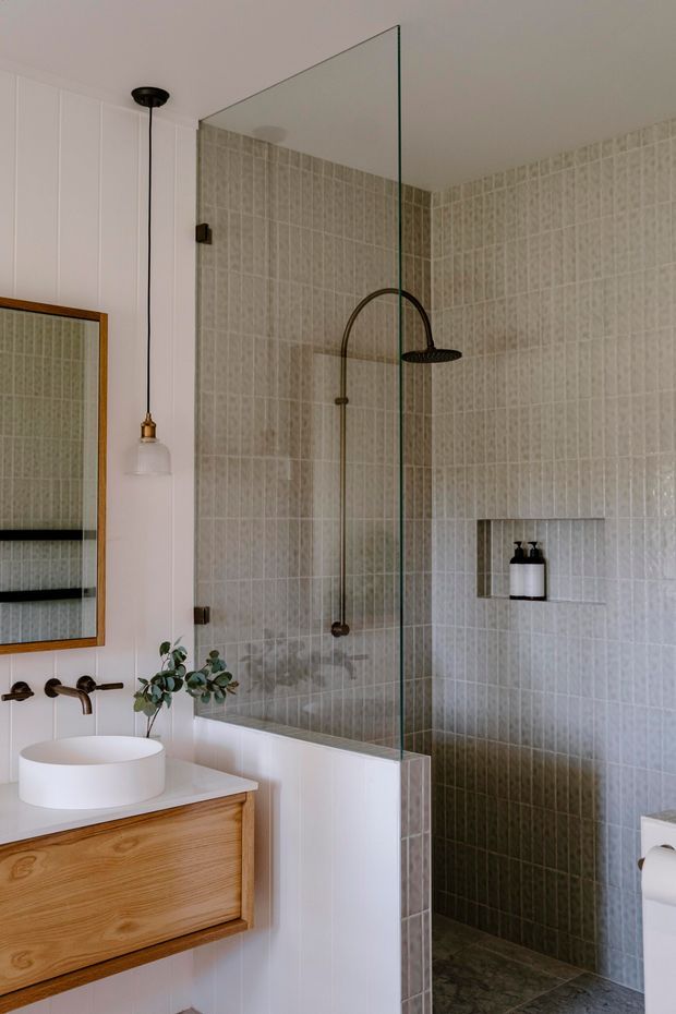 A compact, warmly styled bathroom with a walk-in shower enclosed by a clear frameless glass screen. The shower and adjacent walls are finished in small pale grey-beige mosaic tiles, with a dark bronze or black arched shower fixture and recessed shelf. A timber vanity with a white vessel basin sits against white vertical wall panelling, beside a framed mirror and pendant light. The space has a refined, natural, contemporary feel.