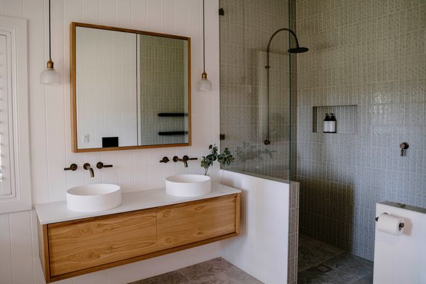 A refined bathroom with a double floating timber vanity, two white vessel basins, wall-mounted bronze-toned tapware, and a large timber-framed mirror. The shower area is enclosed by frameless glass and finished in small pale green-grey mosaic tiles, with a curved dark shower arm and recessed niche. Vertical white wall panelling and pendant lights add a classic architectural character.