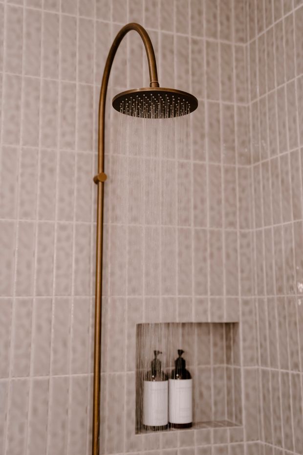 Close-up view of a bathroom shower featuring a brushed bronze or brass rainfall showerhead and vertical riser against pale pink-beige textured square wall tiles. A recessed tiled niche holds two white toiletry bottles.