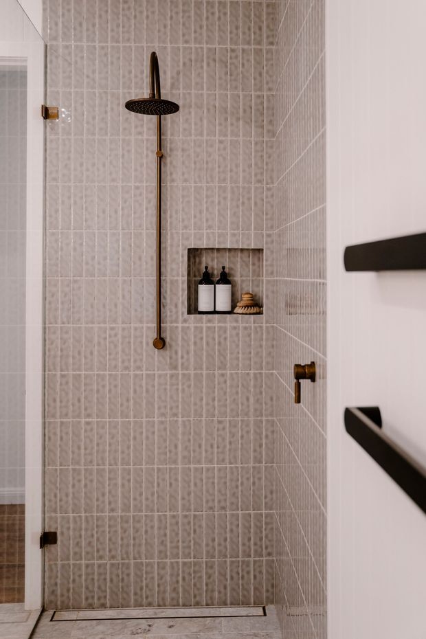 A compact, refined walk-in shower with full-height vertically arranged light grey textured tile walls, a matching tiled floor, and a recessed niche containing dark bottles and a small brush. The shower features a round rainfall showerhead, hand shower rail, and controls in an aged brass or bronze finish, with a frameless glass door and dark hardware visible at the right edge.