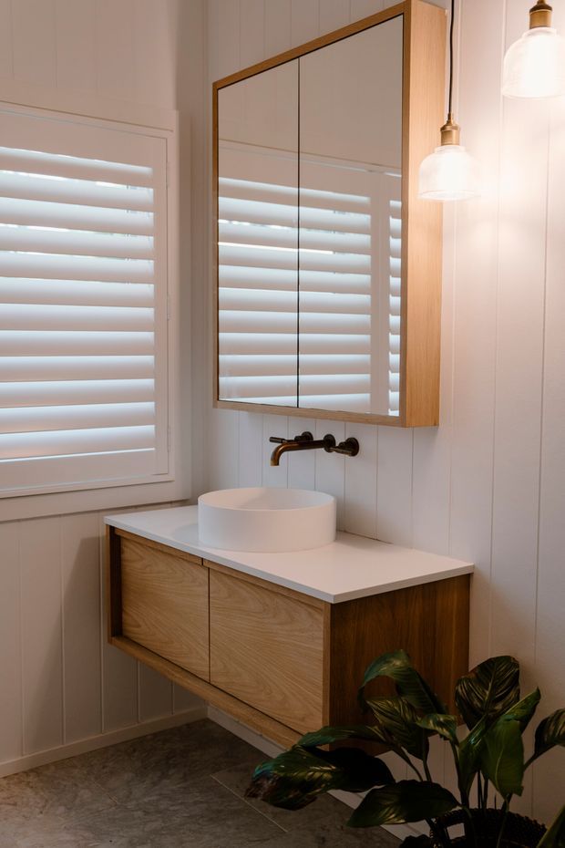 A compact, warmly lit bathroom featuring a floating timber vanity with white benchtop, round vessel basin, wall-mounted bronze tapware, mirrored timber-framed storage cabinet, white vertical wall panelling, plantation shutters, pendant lights, and dark stone-look floor tiles. A leafy indoor plant occupies the foreground.