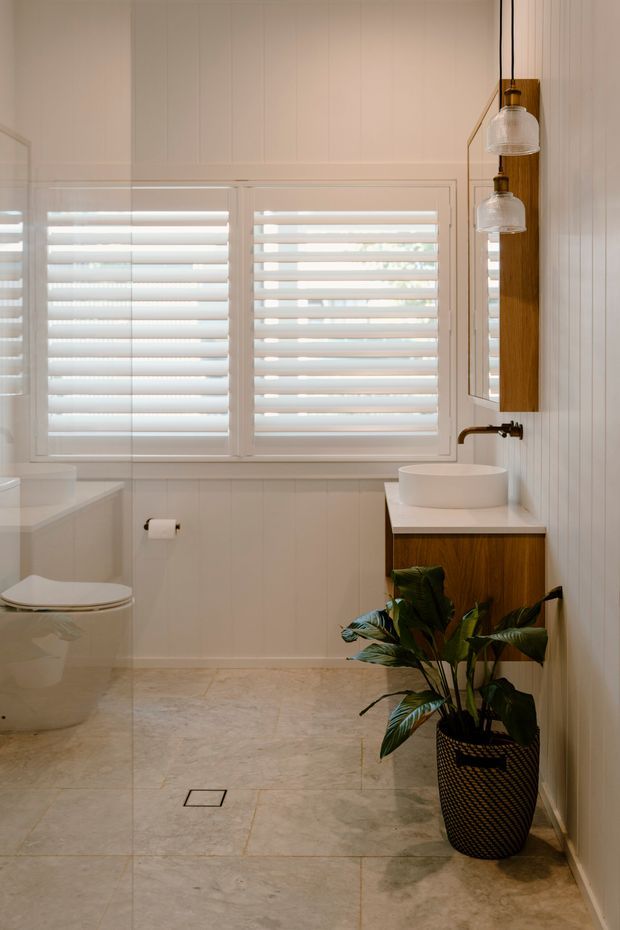 Bright, compact contemporary bathroom with white vertical wall panelling, large white plantation-shutter windows, pale stone-look tiled flooring, a wall-mounted timber vanity with a white vessel basin, brass-toned tapware, pendant lights, and a toilet. A tall potted plant adds greenery.