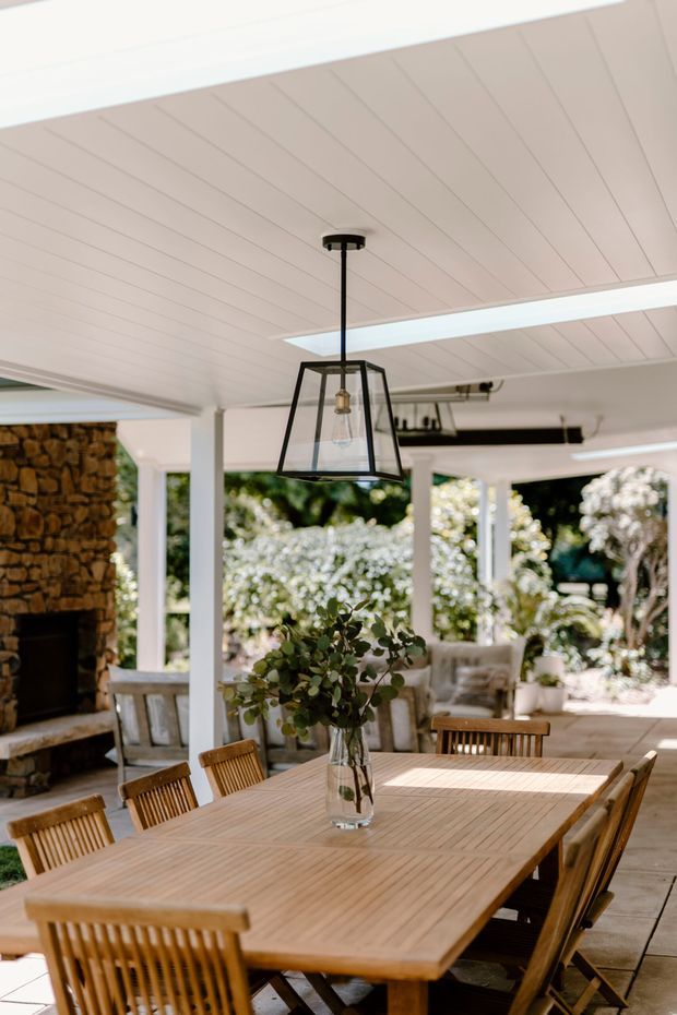 A bright, covered outdoor dining area with a large timber dining table and matching slatted timber chairs. The space features a white paneled ceiling with long skylight-like openings, a black-framed hanging lantern, white structural posts, a substantial natural stone fireplace at the left, and views of lush garden planting beyond.