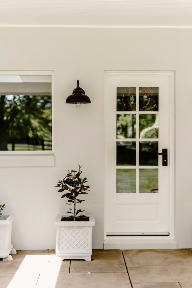 Bright residential entry porch featuring a white paneled exterior door with divided-light glazing, black lever hardware, white decorative trim, a black wall-mounted lantern, and a potted ornamental plant. A matching window and garden reflections are visible, with a paved concrete floor and clean traditional detailing.