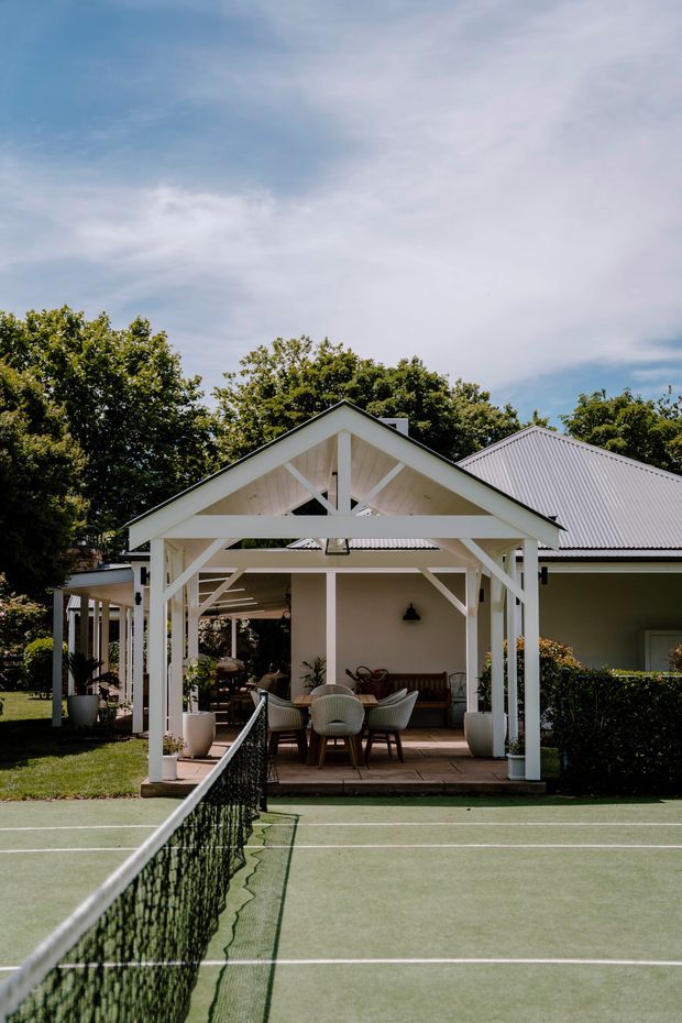 Exterior view of a white-painted gabled pavilion and covered outdoor entertaining area beside a green tennis court. The pavilion has exposed timber framing, a corrugated metal roof, patio seating and dining furniture, with a larger matching building behind it and mature trees surrounding the property.
