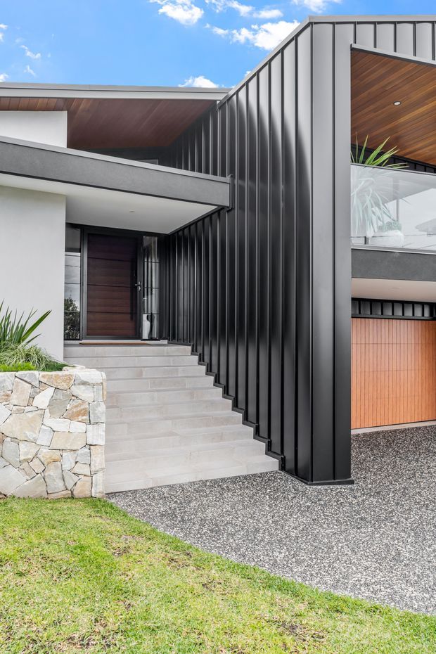 Street-facing view of a contemporary multi-level residence with a dramatic black standing-seam metal-clad facade, broad concrete entry stairs, a dark timber front door, warm timber soffits and balcony ceilings, a glazed balcony, and a timber garage door. A rough natural-stone retaining wall and landscaped lawn frame the entry.