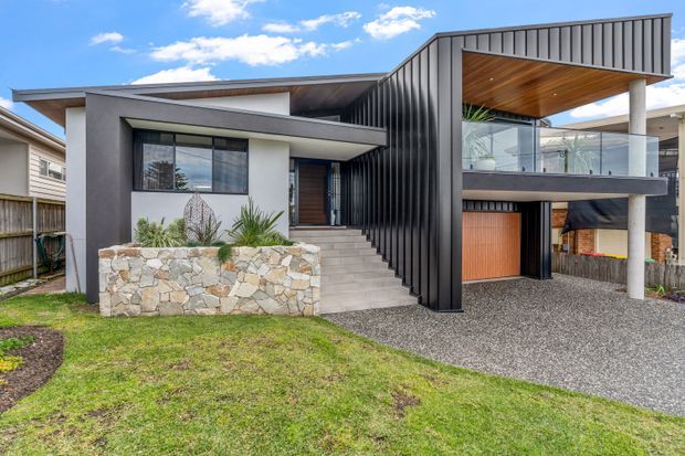 Exterior view of a contemporary multi-level residence with a sharply angled roofline, dark vertical metal cladding, white rendered walls, and exposed concrete columns. A raised balcony with glass balustrading and a timber-lined soffit sits above a garage, while a stone retaining planter and gravel driveway frame the front entrance.