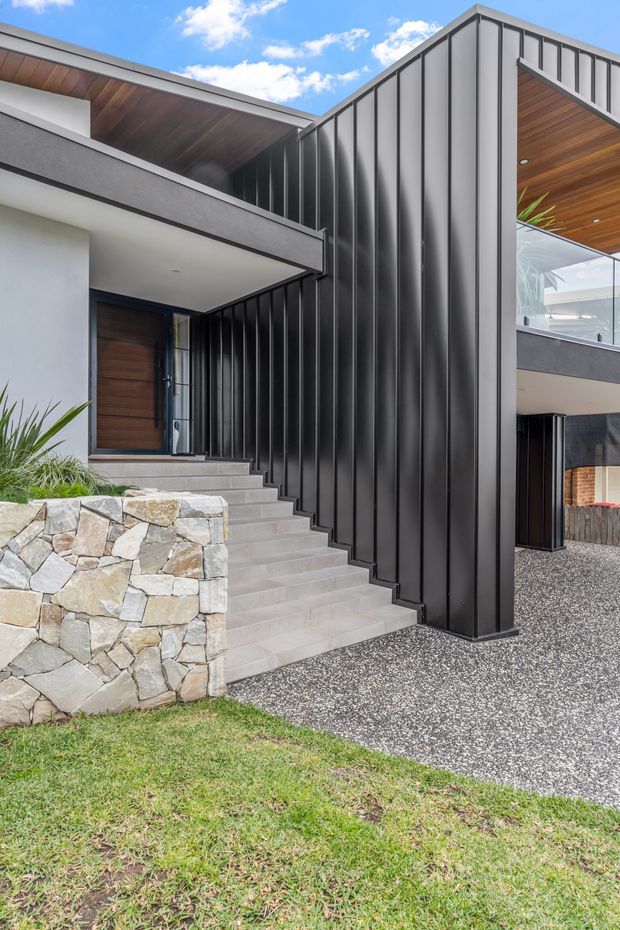 Contemporary multi-level house exterior viewed from the front, featuring dramatic black vertical metal cladding, crisp geometric rooflines, warm timber soffits, a timber entry door, broad concrete steps, and a natural irregular-stone retaining wall. A gravel driveway and lawn occupy the foreground, with glimpses of glazed and covered outdoor areas to the right.