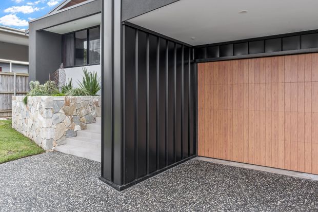 Contemporary residential exterior showing a covered carport or garage entrance with a large timber garage door, black vertical metal cladding and trim, and a contrasting natural stone planter wall with drought-tolerant planting. The textured dark driveway and clean rectilinear form reinforce the modern design.