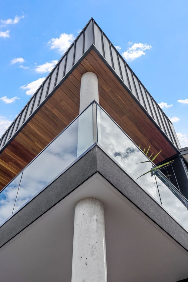 Low-angle exterior architectural photograph of a sharply peaked, multi-level modern building. The facade combines dark metal panel cladding, warm timber soffits, expansive reflective glazing with dark frames, smooth white concrete columns, and a projecting balcony with a glass balustrade. Bright blue sky and clouds are visible around the building.