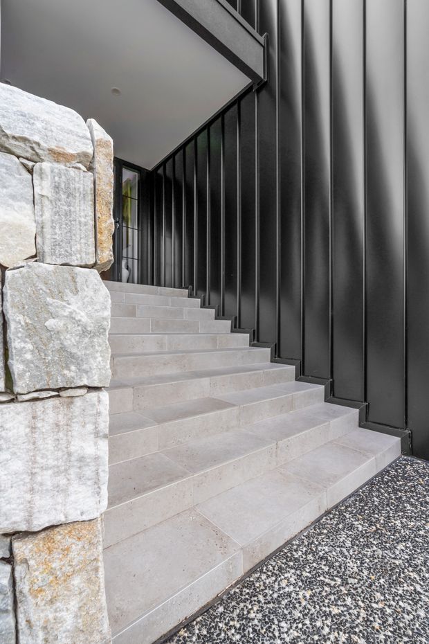 Architectural view of a contemporary exterior stairway leading to a covered entry. The stairs are finished in pale stone or concrete tile, bordered by a rugged stacked-stone wall on one side and a tall black vertically ribbed metal-clad wall on the other. Dark-framed glazing is visible at the upper landing, while a speckled aggregate paving surface occupies the foreground.