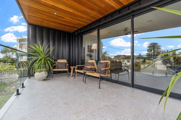 A covered outdoor terrace or balcony with a polished concrete floor, glass balustrade, black-framed sliding glass doors, and a dramatic black vertical-panel wall. Warm timber soffit lining overhead contrasts with the dark exterior cladding. Several chairs and small tables create an outdoor seating area, with indoor living space visible through the glazing and landscaped suburban surroundings beyond.