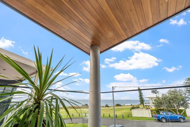 View from a covered exterior terrace or balcony beneath a warm timber-lined soffit, supported by a round concrete column. The image overlooks a suburban or coastal landscape with lawn, roadway, neighbouring homes, and distant water or horizon under a bright blue sky.