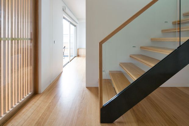 Bright minimalist interior hallway with warm timber flooring and a vertical timber slat screen on the left. A contemporary staircase with open timber treads, a timber handrail, and a bold black structural balustrade dominates the right side. White walls and glazing create an airy, clean-lined space.