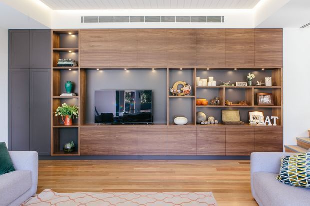 Contemporary living room featuring a full-height custom timber entertainment and storage wall, integrated television, open display shelving, recessed lighting, and dark cabinetry. Warm timber flooring and upholstered seating create a polished, inviting interior.