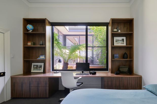 A refined bedroom with an integrated home office along the window wall. Dark timber built-in shelving and cabinetry frame a large black-framed window overlooking a leafy courtyard, with a desk, computer, and white swivel chair beneath. A bed with pale blue bedding occupies the foreground, and ornate crown moulding adds a traditional architectural detail.