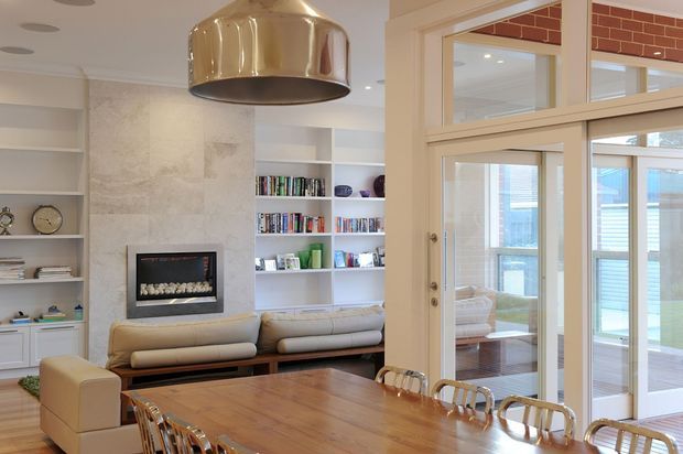 Bright open-plan living and dining area with a long timber dining table and transparent dining chairs in the foreground. The living zone includes a large neutral sectional sofa, built-in white shelving filled with books and objects, and a contemporary inset fireplace set into a pale textured feature wall. A large metallic pendant light hangs above the dining table. Glazed doors and internal windows open toward a bright outdoor or enclosed terrace area, with a small exposed red-brick detail visible above the glazing.