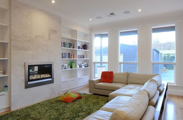 Bright, spacious living room with a large cream sectional sofa arranged around a contemporary built-in fireplace set within a full-height pale stone feature wall. White built-in shelving and cabinetry display books and decor, while tall windows and a glazed door provide abundant natural light. A textured green rug and orange cushions add colour to the otherwise light neutral interior.