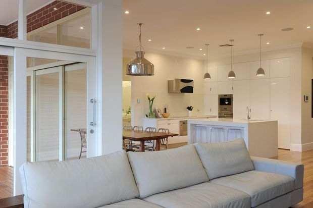 A bright open-plan living, dining and kitchen interior viewed from the living area. A large light-grey sectional sofa dominates the foreground, with a timber dining table and chairs beyond. The kitchen features extensive white full-height cabinetry, a pale island bench, integrated appliances and pendant lighting. White-framed glazed internal doors and windows reveal exposed red brick wall surfaces, adding contrast to the otherwise light, contemporary space.