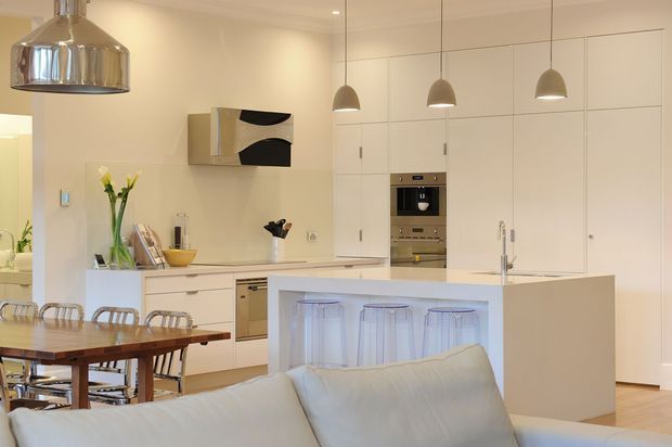 A bright, open-plan contemporary kitchen and dining area viewed from a living room. The kitchen features extensive white flat-panel cabinetry, integrated appliances, a pale island with a raised breakfast counter, stainless-steel pendant lights, and a timber dining table with metal-framed chairs. A white sofa occupies the foreground, creating a connection between the living and kitchen zones.