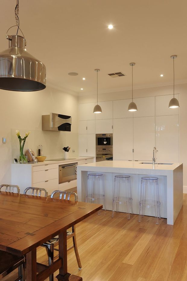 Bright contemporary kitchen and dining area with white full-height cabinetry, integrated stainless-steel appliances, a central island with waterfall-style white benchtop and clear bar stools, pendant lights, and warm timber flooring. A large timber dining table occupies the foreground.
