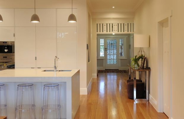 A bright, elongated open-plan kitchen and hallway in a traditional-style home. The kitchen features glossy white full-height cabinetry, a white island with integrated sink and seating for three, stainless-steel tapware, and pendant lights. Polished warm timber flooring continues through the hallway to a glazed entry door, with a console table and stools along the wall. Decorative ceiling details and a transom-style screen above the entry reinforce the classic architectural character.