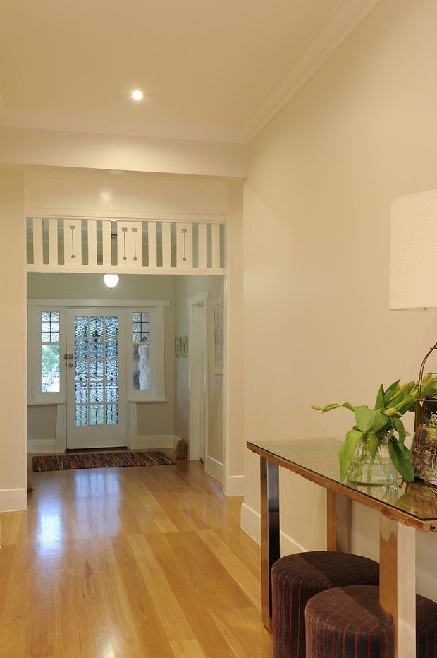 A warm, traditional residential entry hall with polished timber flooring, cream-painted walls and ornate ceiling cornices. The view looks toward a glazed front door with sidelights and a colorful runner, beneath an upper-level balustrade overlooking the hall. A narrow console table with upholstered stools and a table lamp occupies the right side, while internal doors line the corridor.