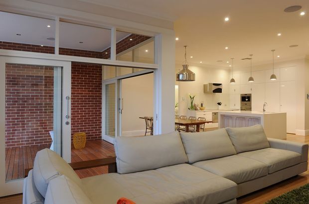 Open-plan living room with a large light-grey sectional sofa in the foreground, looking toward a bright white kitchen and adjacent dining area. Full-height internal glazing and doors frame a brick-lined courtyard or enclosed outdoor space with timber decking, while recessed ceiling lights and pendant fixtures illuminate the room. The kitchen features white floor-to-ceiling cabinetry, a central island with a light timber-toned panelled base, and integrated appliances.